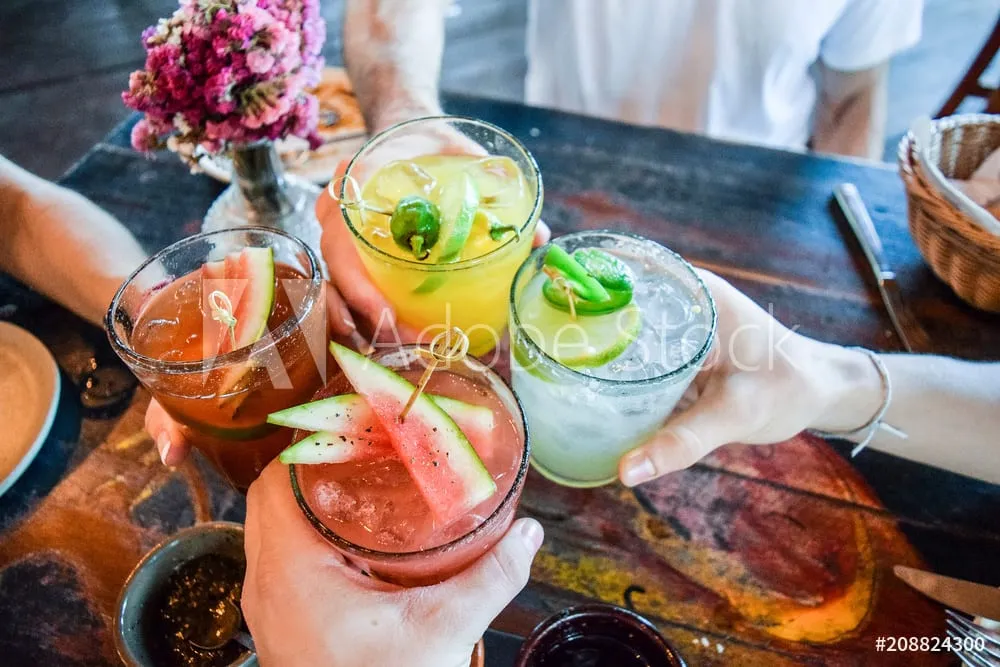 Friends clinking colorful cocktails over a wooden dinner table
