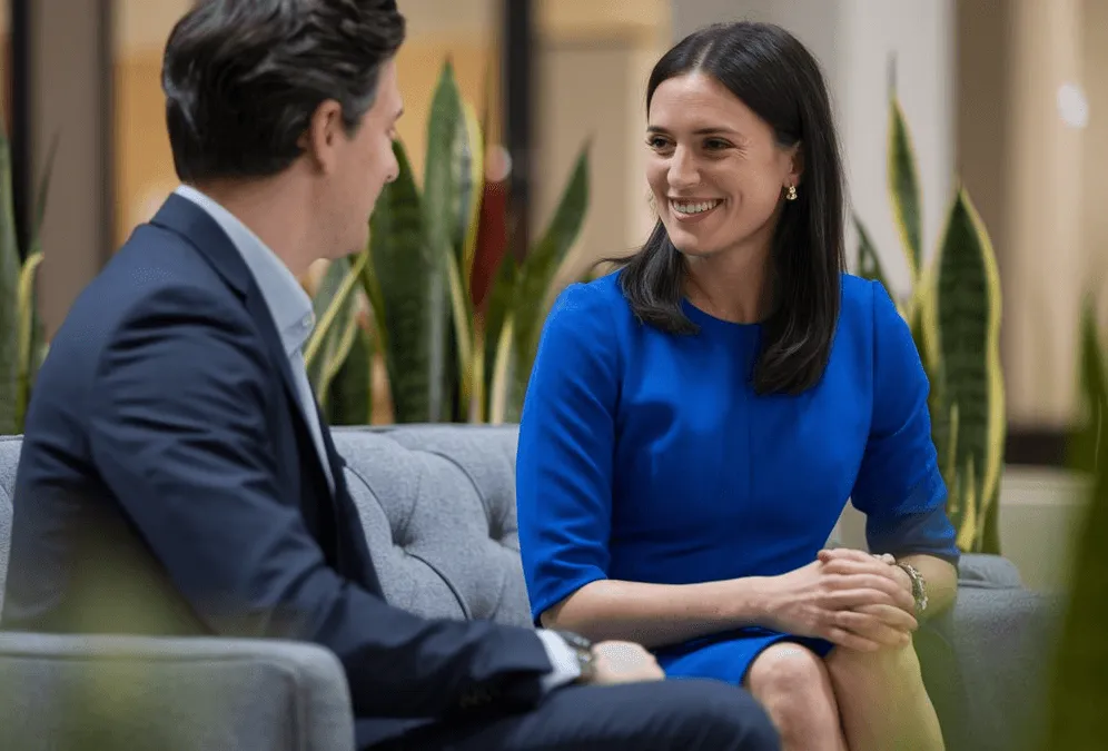 A man and a woman in business attire smiling at each other across a couch